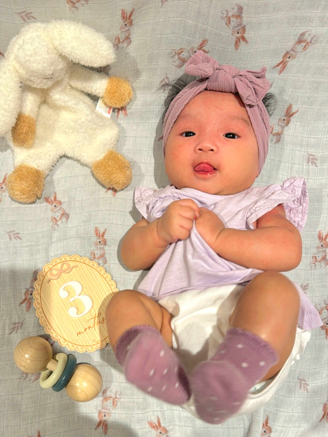 Liara is registered to the contest to win money with this photo: baby, infant, headband, socks, blanket, stuffed_animal, toy, rattle, milestone_card, lying_down, tongue_out, cute, pink, purple, wooden_toy, bunny_pattern, child, soft_toy, playful, clothing