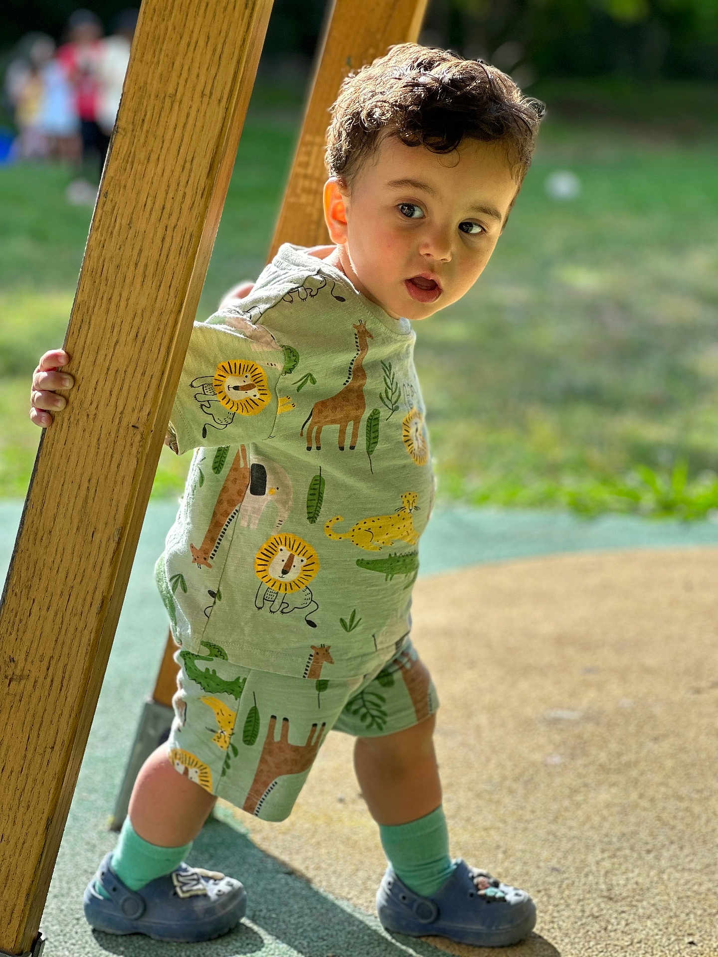 Matheo a rejoint le concours — aidez-le/la à gagner de superbes lots ! toddler, child, outdoor, playground, wood, post, animal_print, shorts, shirt, curly_hair, socks, shoes, greenery, daylight, standing, expression, face, person, summer, playing