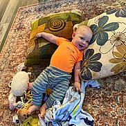 Grady joined the competition — help win amazing prizes! baby, blanket, child, comfort, cozy, cute, face, floor, happy, indoors, laying_down, orange_shirt, person, pillows, playtime, plush_toys, rug, smiling, striped_pants, young_child