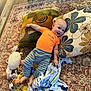 baby, blanket, child, comfort, cozy, cute, face, floor, happy, indoors, laying_down, orange_shirt, person, pillows, playtime, plush_toys, rug, smiling, striped_pants, young_child