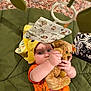 baby, teddy_bear, orange_clothing, green_cushion, yellow_towel, baby_toy, playful, child, cute, indoor, soft_toy, mobile, lying_down, infant, cozy, person, plush, face, hand, curious
