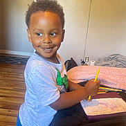 Amir is registered to the contest to win money with this photo: child, smiling, crayon, drawing, notebook, scribbles, indoor, wooden_floor, blanket, table, clothing, curly_hair, person, happy, art, colorful, casual, home, young, creative