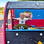 bench, blonde_hair, blue_sky, child, daylight, fence, fun, grass, happy, outdoor, park, play_structure, playground, recreation, short_sleeve_shirt, smiling, steering_wheel, toddler, toy_vehicle, trees