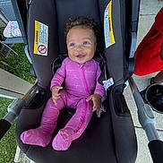 Eilena Lea joined the competition — help win amazing prizes! baby, car_seat, pink_onesie, smiling, curly_hair, blue_eyes, infant, outdoor, grass, sidewalk, person, happy, clothing, footed_pajamas, child, seatbelt, fabric, hand, portrait, cute