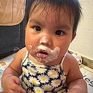 Catalina is registered to the contest to win money with this photo: baby, child, face, cream, yogurt, messy, daisy_pattern, clothing, indoor, curious, hair, sitting, play_mat, skin, person, cute, portrait, infant, young_child, expression
