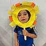 child, toddler, craft, paper_plate, sunflower_mask, yellow_color, orange_color, crepe_paper, wooden_stick, blue_shirt, sports_shirt, striped_sleeves, wall_background, indoor_scene, face, eyes, hand, portrait, smile, classroom_activity