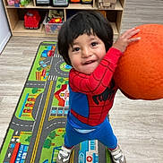 Gino is registered to the contest to win money with this photo: child, toddler, playroom, toy_shelf, playmat, orange_ball, spiderman_costume, smile, standing, sneakers, wooden_floor, toy_truck, blocks, rug, cabinet, indoor, young_child, hair, hands, portrait