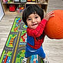 child, toddler, playroom, toy_shelf, playmat, orange_ball, spiderman_costume, smile, standing, sneakers, wooden_floor, toy_truck, blocks, rug, cabinet, indoor, young_child, hair, hands, portrait