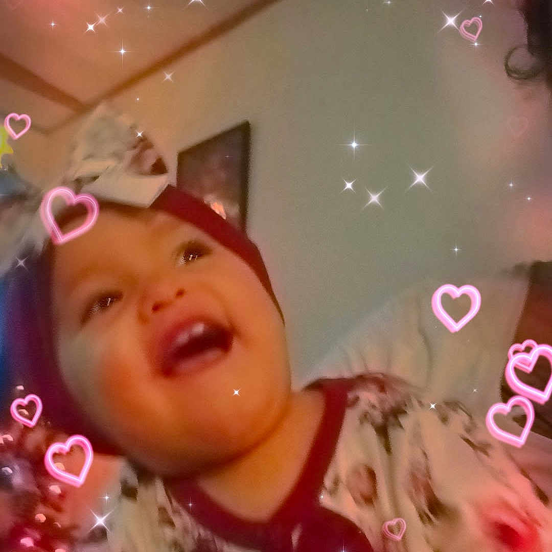 Gena is registered to the contest to win money with this photo: baby, child, headband, bow, floral_clothing, smiling, happy, indoor, person, sparkles, hearts, christmas_tree, holiday, portrait, face, cute, joy, celebration, infant, warm_lighting