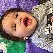 James is registered to the contest to win money with this photo: child, toddler, smile, open_mouth, teeth, laughing, face, hair, eyes, grey_shirt, basketball_graphic, play_mat, purple_background, green_background, textured_mat, portrait, indoors, happy, casual_clothing, baby