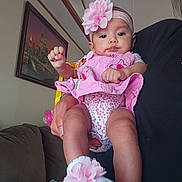 Gena is registered to the contest to win money with this photo: baby, infant, pink, flower_headband, flower_socks, onesie, person, indoor, living_room, sofa, hand, portrait, cute, child, accessory, decoration, clothing, wall_art, clock, holding
