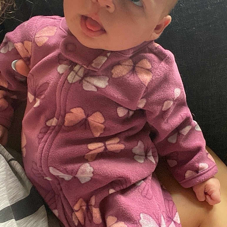 Ariyanna joined the competition — help win amazing prizes! adult_arm, baby, butterfly_pattern, casual, child, clothing, couch, curious_expression, face, head, holding, indoor, infant, person, purple_onesie, seated, skin, soft_fabric, tongue_out, window