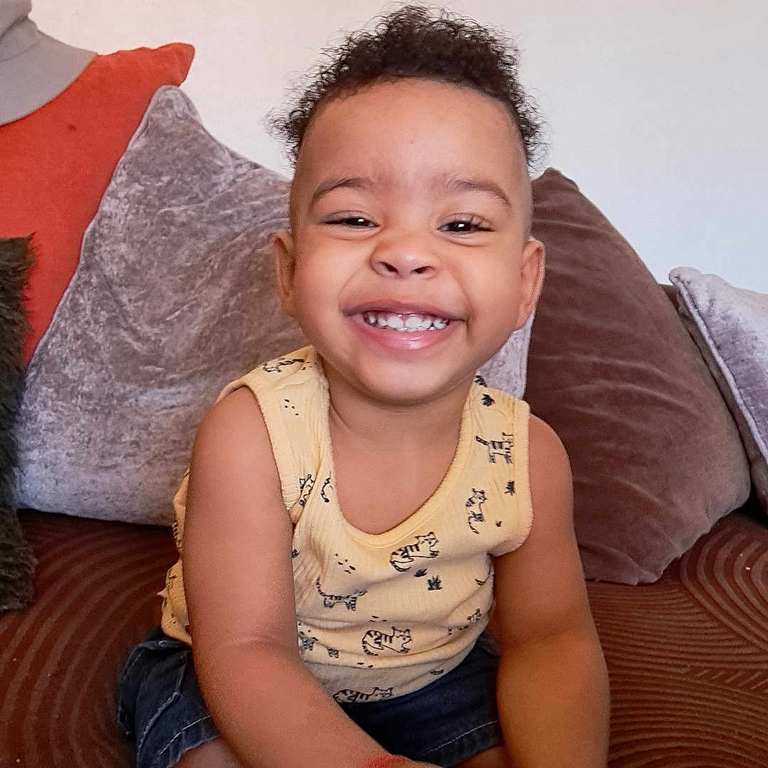 Caelen a rejoint le concours — aidez-le/la à gagner de superbes lots ! child, couch, cozy, curly_hair, cute, denim_shorts, footwear, hand, happy, home, indoor, person, pillows, playful, sitting, skin, sleeveless_shirt, smile, toddler, yellow_shirt