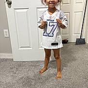 Marielys is registered to the contest to win money with this photo: barefoot, broom, buffalo, carpet, casual, child, clothing, door, door_handle, floor, football_jersey, hair, indoor, person, room, smiling, standing, wall_outlet, white_door, young_child