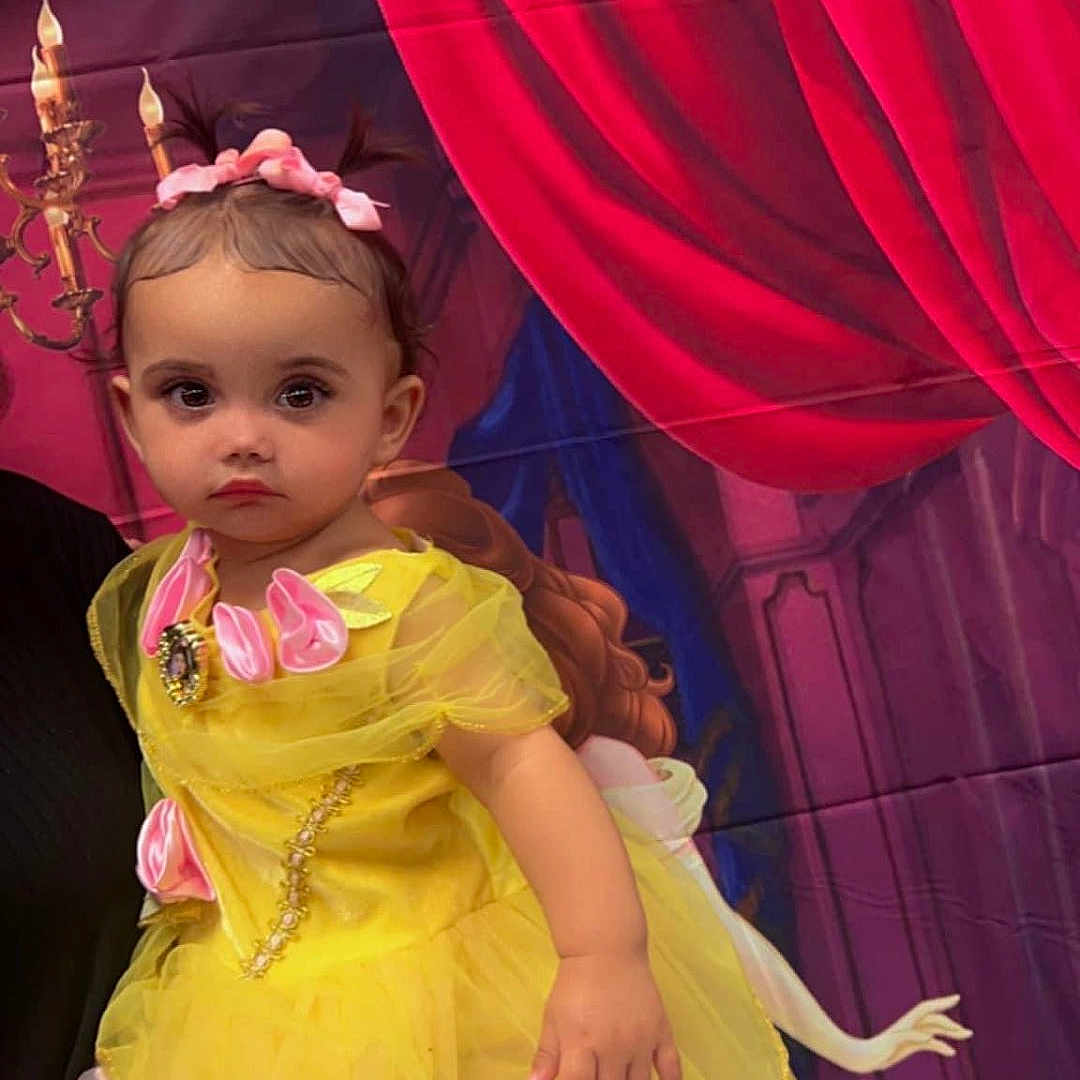Marielys is registered to the contest to win money with this photo: baby, celebration, child, costume, curtains, cute, dress, face, hand, holding, indoor, party, person, pink_bows, portrait, princess_costume, serious_expression, theater_background, toddler, yellow_dress