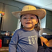 Liam is registered to the contest to win money with this photo: toddler, child, smiling, hat, cowboy_hat, indoor, kitchen, person, striped_shirt, text_on_clothing, happy, face, long_hair, blue_shirt, cabinet, countertop, appliance, casual_clothing, holding, portrait