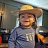 toddler, child, smiling, hat, cowboy_hat, indoor, kitchen, person, striped_shirt, text_on_clothing, happy, face, long_hair, blue_shirt, cabinet, countertop, appliance, casual_clothing, holding, portrait