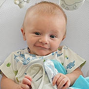 Beckham joined the competition — help win amazing prizes! baby, infant, child, portrait, close_up, smiling, wide_eyed, sitting, onesie, dinosaur_print, blanket, stuffed_toy, hanging_toy, nursery, crib, bedding, indoor, hands, blue, face