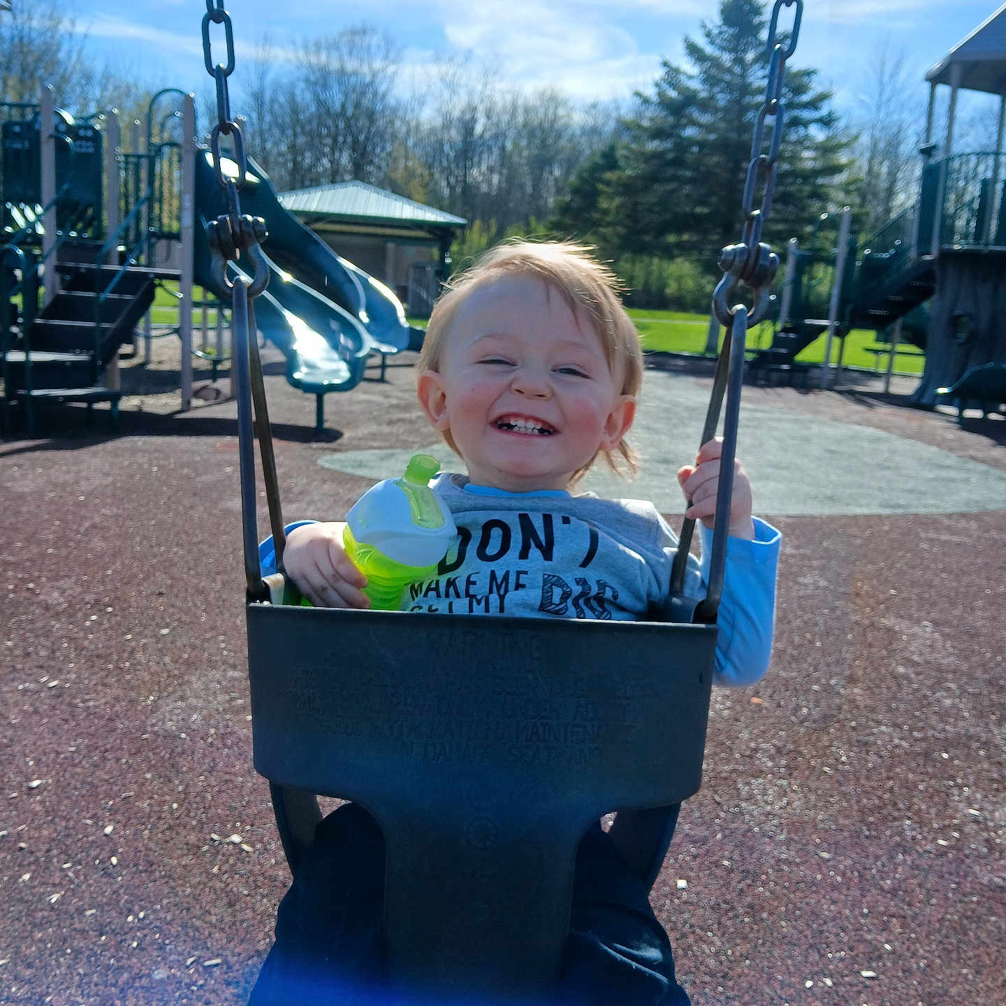 Tobias joined the competition — help win amazing prizes! child, toddler, swing, playground, smiling, happy, outdoor, daylight, park, clothing, holding, green, sippy_cup, sky, trees, slide, play_equipment, fun, joy, person