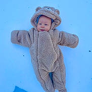 Jace is registered to the contest to win money with this photo: baby, child, snow, winter, onesie, costume, fuzzy, cute, outdoor, cold, face, tongue, blue, warm_clothing, lying_down, playful, headwear, person, infant, animal_ears