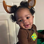 Jakodah is registered to the contest to win money with this photo: toddler, child, smiling, face, curly_hair, brown_clothing, reindeer_ears, indoor, person, holding, door, cute, happy, portrait, baby, costume, accessory, black_hair, hand, playful