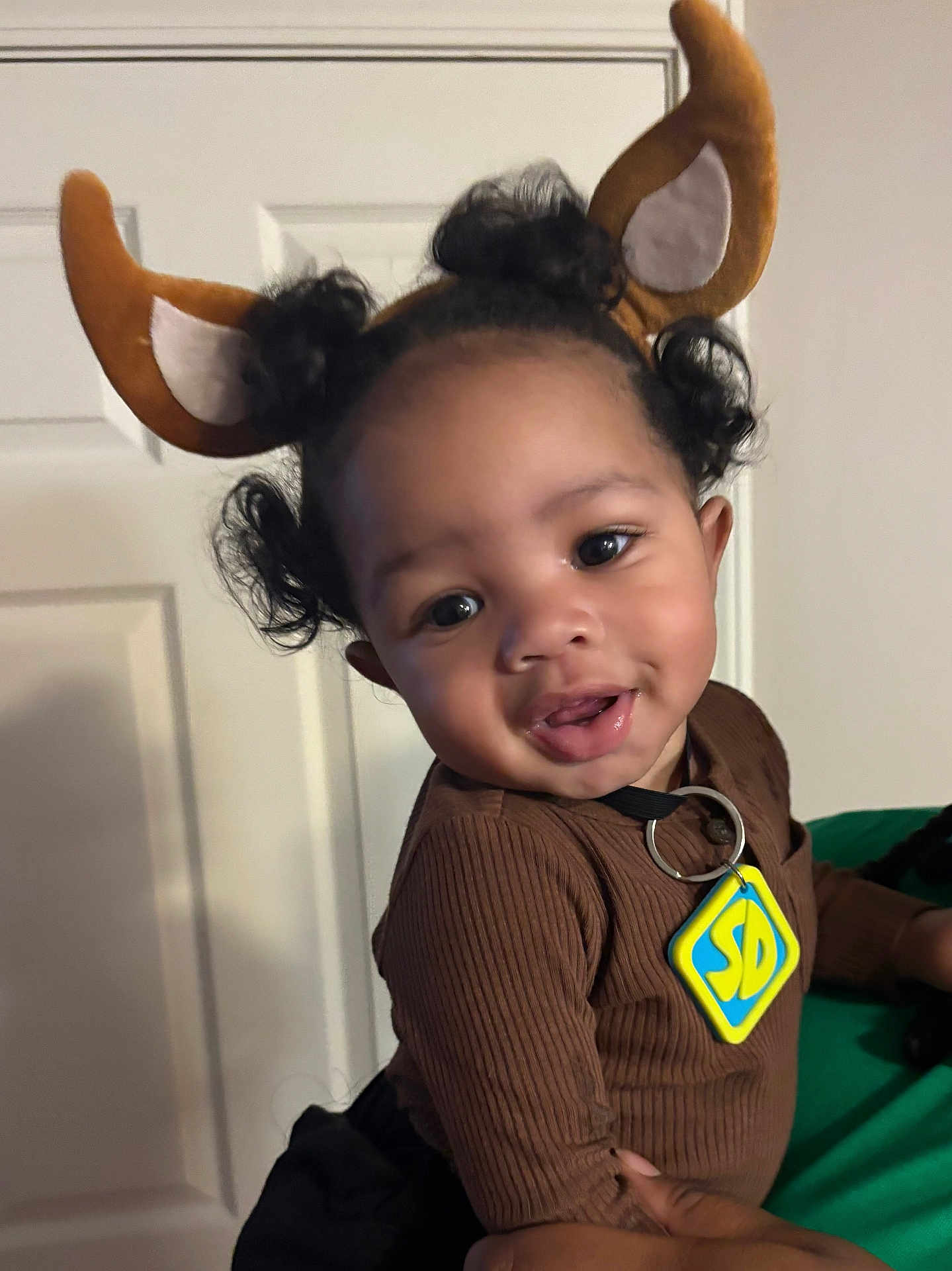 Jakodah is registered to the contest to win money with this photo: toddler, child, smiling, face, curly_hair, brown_clothing, reindeer_ears, indoor, person, holding, door, cute, happy, portrait, baby, costume, accessory, black_hair, hand, playful