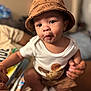 baby, child, hat, bucket_hat, white_shirt, basketball_graphic, holding_hand, person, indoor, cute, face, eyes, pout, skin, sitting, adult_hand, focus, blurred_background, infant, clothing
