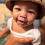 baby, child, smile, hat, brown_hat, white_clothing, hand, person, face, closeup, joyful, cute, indoor, portrait, skin, eyes, infant, young_child, happy, blurred_background