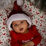 Avianna joined the competition — help win amazing prizes! baby, child, santa_hat, christmas, holiday, festive, red_clothing, blanket, reindeer_pattern, ornament, tree, indoors, cute, infant, portrait, holiday_season, smiling, hands, face, cozy
