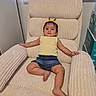 child, toddler, armchair, yellow_bow, yellow_top, denim_shorts, indoor, plush_furniture, relaxed, baby_supplies, window_blind, light_wall, curious_expression, barefoot, home_interior, person, seated, cozy, casual_clothing, portrait