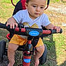 Brittany is registered to the contest to win money with this photo: toddler, child, tricycle, outdoor, grass, sunlight, headband, bow, shorts, white_shirt, gold_heart, barefoot, hand_grip, red_tricycle, black_tricycle, serious_expression, play, summer, toy, young_child