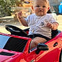 baby, blurred_background, child, cute, face, garden, happy, necklace, outdoor, playing, portrait, red_car, seatbelt, short_sleeves, smiling, steering_wheel, sunlight, toy_car, wheel, white_tshirt