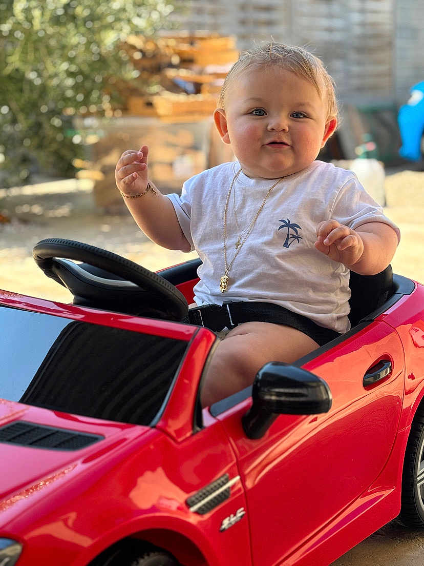 Julio participe au concours pour gagner de l'argent avec cette photo : baby, blurred_background, child, cute, face, garden, happy, necklace, outdoor, playing, portrait, red_car, seatbelt, short_sleeves, smiling, steering_wheel, sunlight, toy_car, wheel, white_tshirt