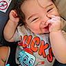 baby, child, infant, portrait, smiling, happy, playful, face, eyes, curly_hair, hands, nose_picking, gray_shirt, cartoon_shirt, car_seat, vehicle, safety_warning, close_up, indoor, colorful