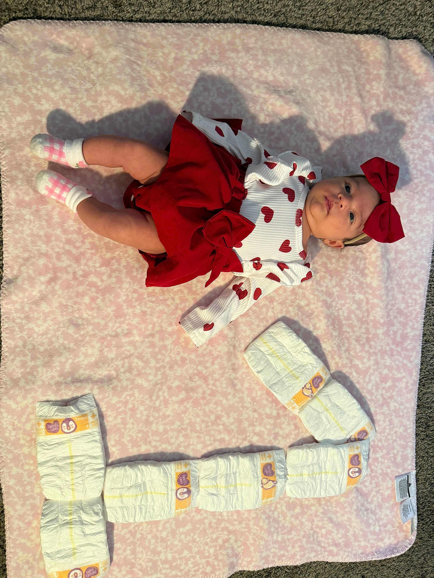 Itzel joined the competition — help win amazing prizes! baby, infant, diapers, blanket, red_bow, heart_pattern, pink, white, socks, cute, laying_down, soft_texture, floor, carpet, child, smile_shape, clothing, headband, playful, indoor