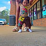 toddler, child, walking, adult_hands, sidewalk, brick_building, outdoor, colorful_clothing, socks, sandals, storefront, signage, car, trees, daylight, person, support, early_steps, urban, casual