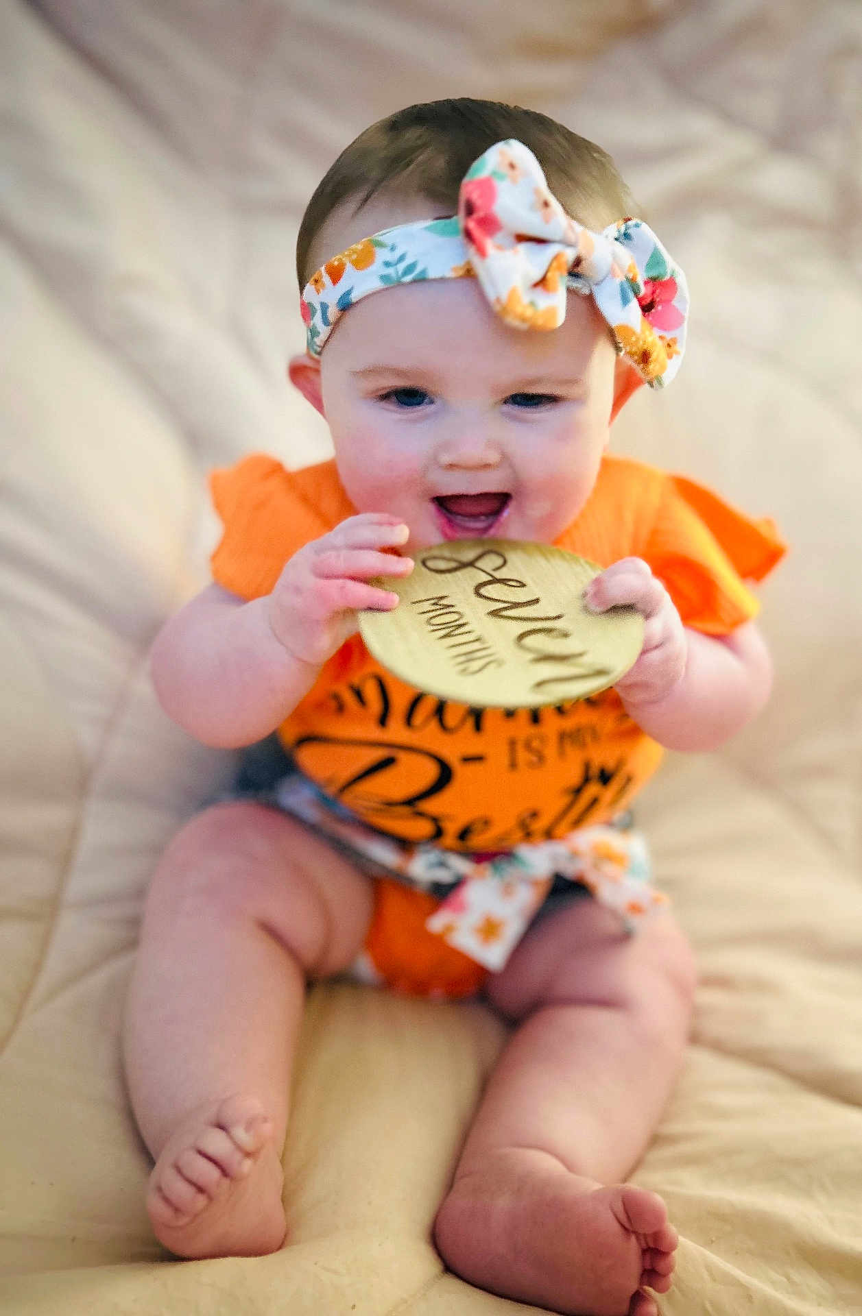 Ezlynn is registered to the contest to win money with this photo: baby, infant, child, orange_clothing, headband, floral_pattern, wooden_plaque, seven_months, milestone, cute, smiling, sitting, barefoot, soft_blanket, indoors, portrait, skin, face, hand, feet