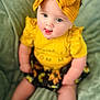 baby, child, yellow_bow, yellow_top, ruffled_sleeves, black_floral_skirt, sitting, tongue_out, blue_eyes, soft_blanket, indoor, cute, portrait, infant, headband, playful, smiling, person, closeup, happy