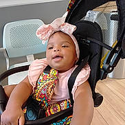 Nicole joined the competition — help win amazing prizes! baby, child, stroller, headband, colorful_clothing, indoor, flooring, chair, smiling, happy, infant, person, seatbelt, pattern, wood_floor, arm, hand, face, cute, toddler
