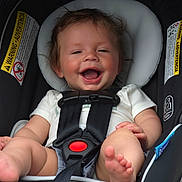 Wyatt is registered to the contest to win money with this photo: baby, infant, car_seat, child, smiling, happy, safety_harness, seatbelt, cute, person, clothing, foot, hand, warning_label, black_straps, white_shirt, shorts, indoors, portrait, closeup
