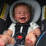 baby, infant, car_seat, child, smiling, happy, safety_harness, seatbelt, cute, person, clothing, foot, hand, warning_label, black_straps, white_shirt, shorts, indoors, portrait, closeup
