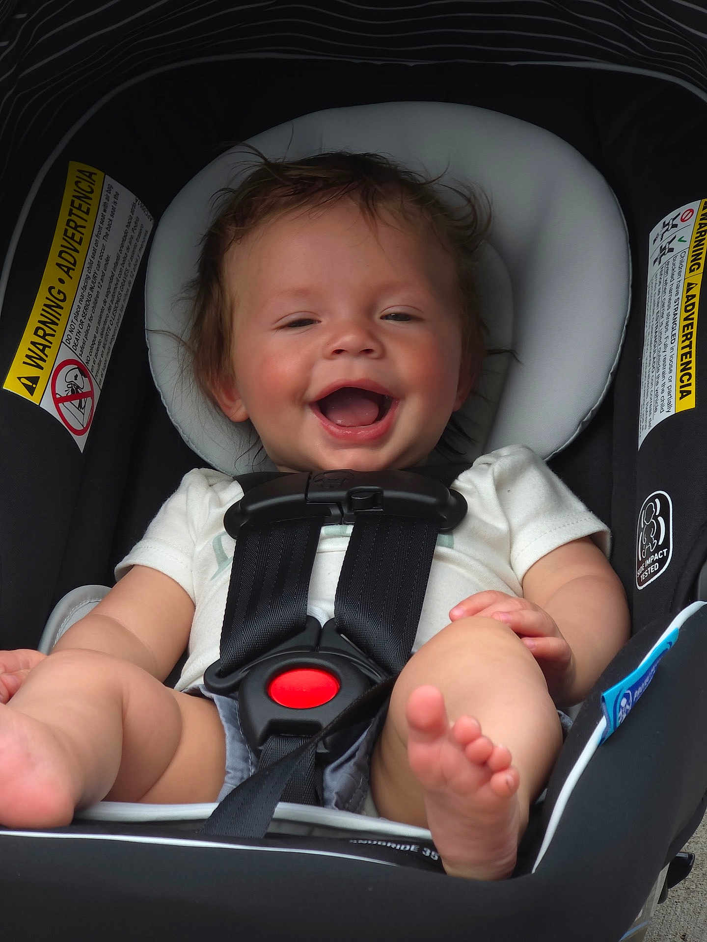 Wyatt is registered to the contest to win money with this photo: baby, infant, car_seat, child, smiling, happy, safety_harness, seatbelt, cute, person, clothing, foot, hand, warning_label, black_straps, white_shirt, shorts, indoors, portrait, closeup