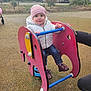 child, toddler, playground, spring_ride, pink_hat, puffer_jacket, jeans, boots, smiling, play_equipment, park, outdoors, trees, building, stroller, adult_hand, daylight, rubber_surface, grass, happy