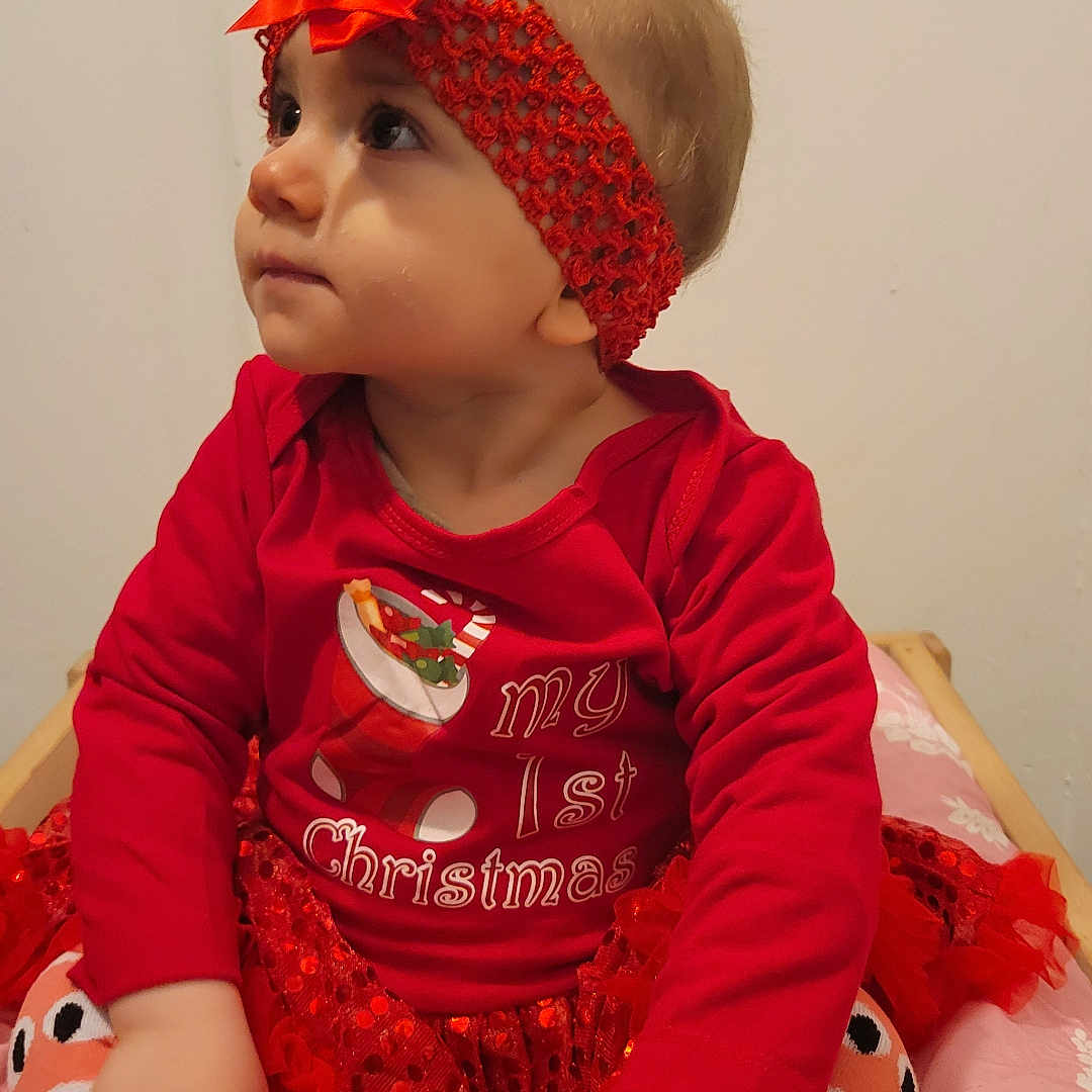 Lilou a rejoint le concours — aidez-le/la à gagner de superbes lots ! baby, child, red_clothing, headband, bow, christmas_theme, festive, sitting, cute, indoor, infant, portrait, holiday, cushion, pattern, looking_up, long_sleeve, toddler, decorative, young_child