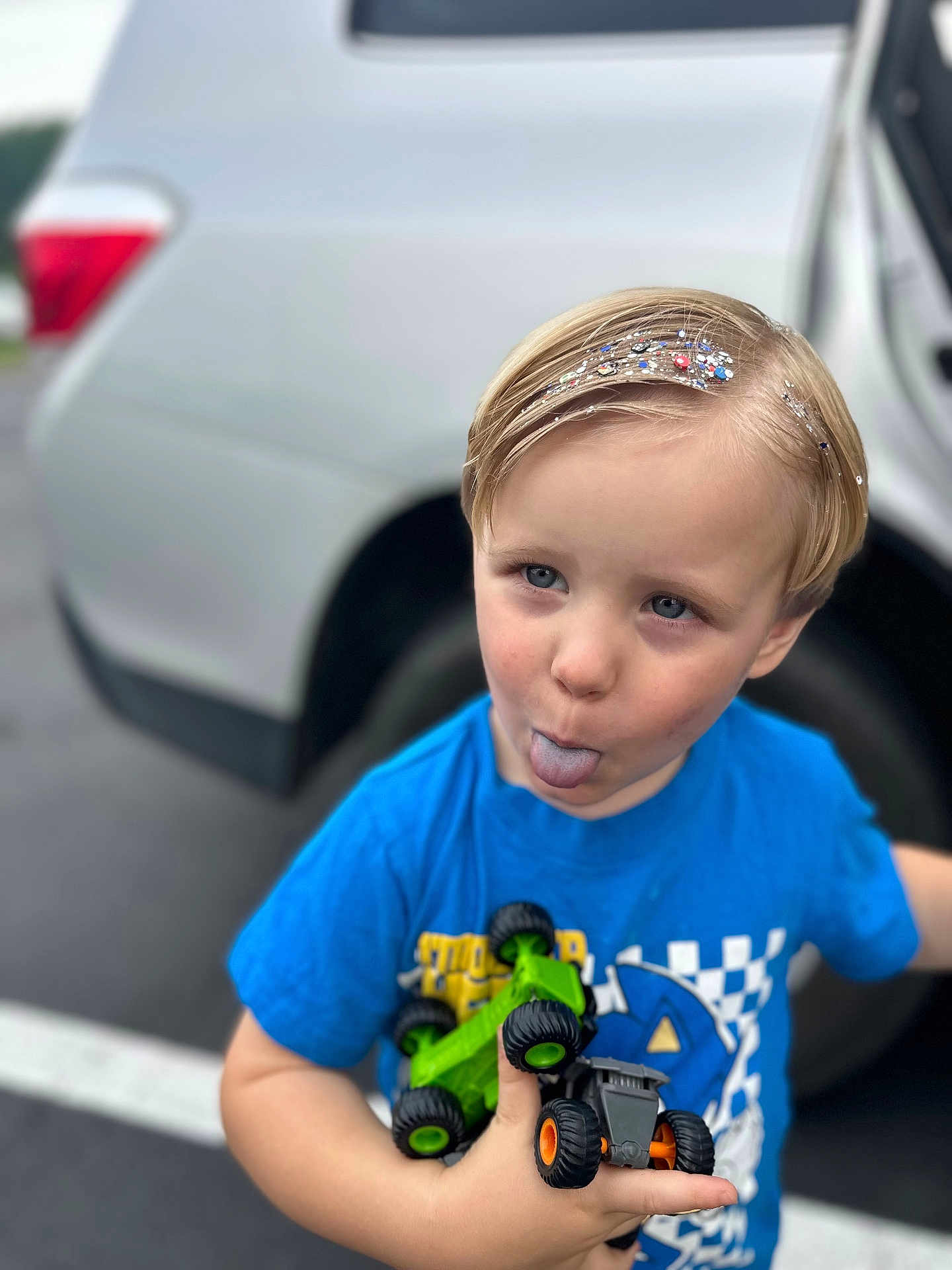Ezekiel is registered to the contest to win money with this photo: child, boy, blue_shirt, toy_trucks, glitter, blonde_hair, playful, parking_lot, car, tongue_out, outdoor, cute, smiling, vehicle, casual_clothing, holding, happy, person, daylight, close_up