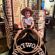 Ezekiel is registered to the contest to win money with this photo: child, boy, cowboy_hat, saddle, lasso, brick_wall, restaurant, sign, indoor, smiling, shorts, tshirt, shoes, wood, decor, rope, sevierville, texas_kitchen, playful, posing