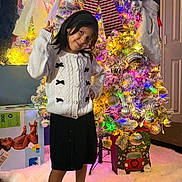 Verónica is registered to the contest to win money with this photo: child, girl, smiling, portrait, christmas, christmas_tree, festive, holiday, ornaments, fairy_lights, santa_hat, winter, sweater, skirt, standing, indoor, home, gifts, colorful, decoration