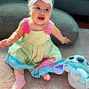 Juana is registered to the contest to win money with this photo: baby, child, infant, smiling, sitting, portrait, headband, bow, dress, plaid, colorful_clothing, pink_socks, plush_toy, stuffed_animal, blue_toy, rug, living_room, couch, indoor, happy