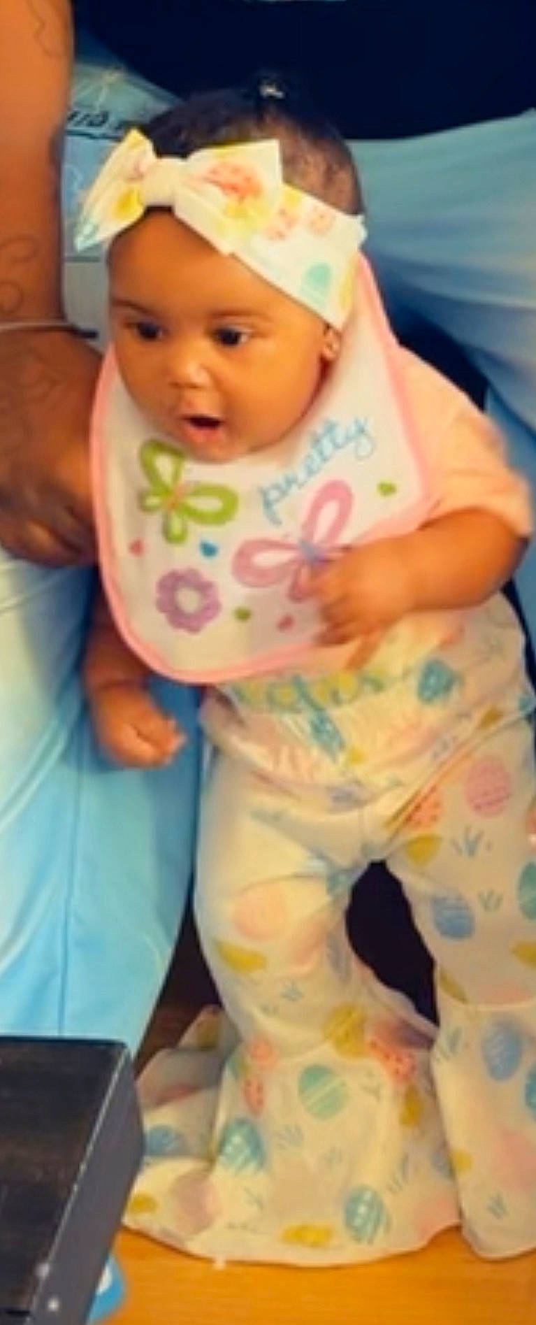 Cailyn is registered to the contest to win money with this photo: adult, baby, bib, bright_colors, child, colorful_clothing, curious, expression, floor, hand, headband, holding, indoor, infant, pants, patterned_clothing, person, standing, support, young_child