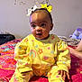baby, child, yellow_outfit, bows, hair_bows, rainbow_pattern, strawberry_pattern, bedspread, colorful, indoor, sitting, curious, person, foot, hand, head, face, expression, wall_decoration, cozy